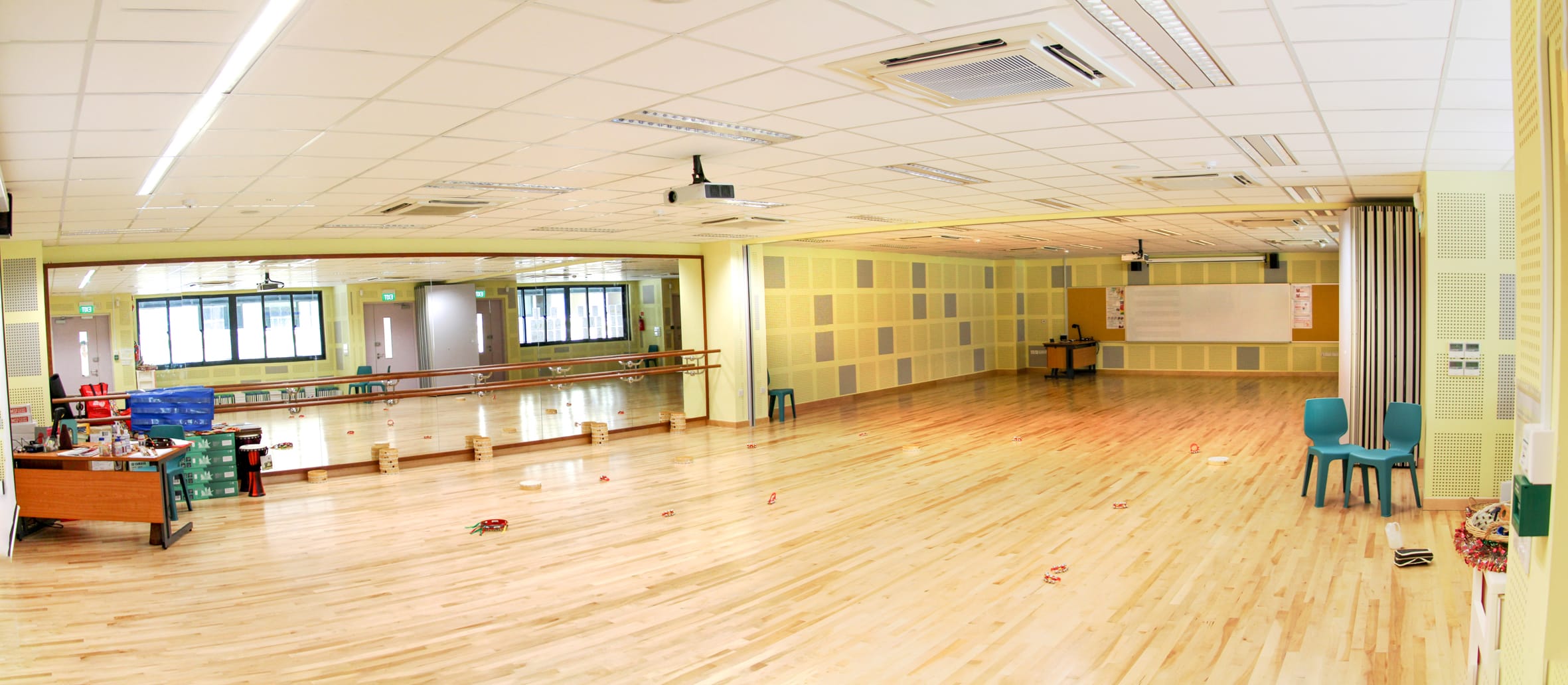 Large empty dance studio with wooden floor, wall mirrors, ballet barre, yellow walls, and teal chairs in the corner.